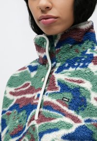 Πουλόβερ από φλίς με ψηλό γιακά, με πράσινα, μπλε και μπουρντό αφηρημένα σχέδια. Περιλαμβάνει λευκές λεπτομέρειες και μια μικρή ετικέτα μάρκας.