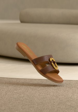 Bruine leren slide-sandaal met gouden gespsluiting, afgebeeld terwijl hij gebogen in de lucht hangt boven een textured beige tapijt en een beige bank op de achtergrond.