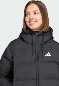 Chaqueta acolchada negra con cuello alto, cierre de cremallera y capucha con cordón. Presenta una textura acolchada sutil y un logo blanco de Adidas.