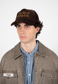 Jeune homme portant une casquette en velours côtelé marron avec le texte "L'art d'être en retard", une chemise rayée bleue et une veste taupe avec un écusson indiquant "Interdit".