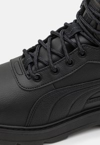 La sneaker haute en cuir noir présente une surface texturée, des lacets solides, des œillets en métal et une semelle en caoutchouc épaisse avec des coutures accentuées.