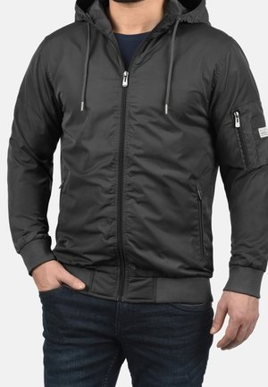 Man wearing dark gray zip-up hooded jacket with drawstrings, zippered pockets on sides and sleeve, paired with dark jeans.