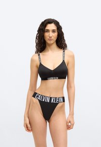 Γυναίκα με σγουρά μαλλιά φοράει μαύρο bralette Calvin Klein και εσώρουχο με ψηλή μέση και ελαστικές ταινίες με λογότυπο, στέκεται μπροστά σε απλό φωτεινό φόντο.