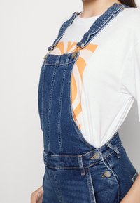 Denim-Latzhose mit verstellbaren Trägern, goldenen Beschlägen, seitlichen Knöpfen und einem lockeren Schnitt. Kombiniert mit einem weißen Grafik-T-Shirt.