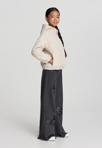 Giacca beige trapuntata con cappuccio, abbinata a pantaloni in denim grigio scuro a gamba larga con stampe grafiche. Le sneaker bianche completano il look.