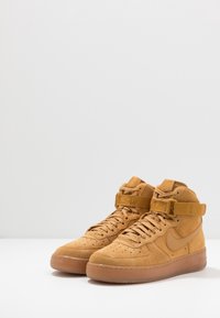 Ett par högklackade sneakers i tanfärgad mocka med snören och ankelremmar, placerade på vit yta mot en ljusgrå bakgrund.