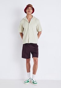 Chemise vert clair à manches courtes avec col, associée à un short marron foncé, des chaussettes blanches et des baskets blanches avec des accents verts. Chapeau seau marron.
