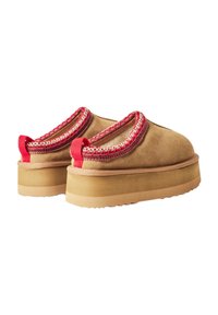 Scarpe slip-on in suede marrone con rifiniture e dettagli decorativi rossi, dotate di suola piatta e suola in gomma testurizzata.
