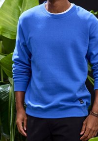 Sweat-shirt bleu en tissu doux avec un col rond, des poignets et un ourlet côtelés, doté d'une étiquette logo noire sur le côté inférieur.