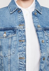 Chaqueta de mezclilla azul con cuello, herrajes plateados, bolsillos frontales y cierre de botones. Llevada sobre una camisa blanca de botones.