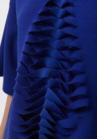 Top azul con volantes en capas y ondulados a lo largo de los lados. Fabricado con una tela suave de textura ligeramente brillante. Manga corta.