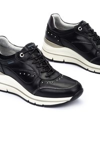 Zapatillas de cuero negro con diseño de cordones, detalles con tachuelas y una suela blanca gruesa. Presentan un acabado suave y un acolchado en el collar para mayor comodidad.
