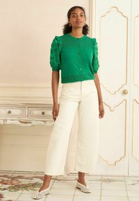 Cardigan verde lavorato a maglia con maniche texturizzate e dettagli volant, abbinato a pantaloni cream a gamba larga e scarpe slip-on bianche traforate.
