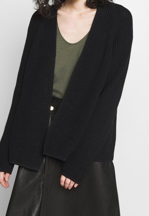 Femme portant un cardigan en grosse maille noire par-dessus un haut vert olive et une jupe en cuir noire avec une ceinture à boutons visible.