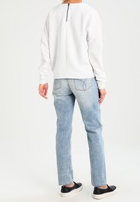 Suéter blanco texturizado con detalle de cremallera en la parte posterior, combinado con jeans de pierna recta azul claro y zapatillas negras tipo slip-on.