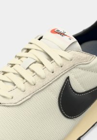 Baskets Nike avec une tige en maille crème, logo noir, semelle en caoutchouc beige et lacets plats. Accents texturés distincts sur le bout et les côtés.