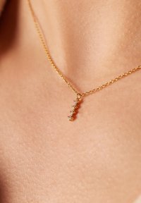 Gouden ketting met een verticale hanger met vijf kleine heldere edelstenen, tegen een huidkleurige achtergrond.