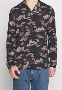 Schwarze Knopfhemd mit rosa und beigen Drachen- und Wolkenmustern, getragen mit blauen Jeans, zeigt den Oberkörper und die Hände.
