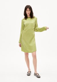Grünes, satinartiges Kleid mit langen Ärmeln und abgerundetem Saum. Das Model trägt schwarze, mit Nieten verzierte Schuhe und steht vor einem weißen Hintergrund.