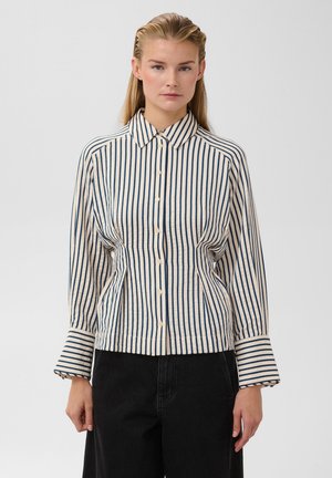 Femme portant une chemise à manches longues boutonnée avec des rayures verticales bleues et blanches et un pantalon noir, debout devant un fond uni.