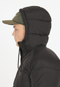Svart vadderad jacka med hög krage, försedd med en dragsko på huvan. Modellen bär en grön keps under huvan. Slät textur.