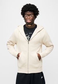 Μπεζ φούτερ με φερμουάρ και απαλή υφή, με μπροστινή τσέπη και κουκ ούλα με κορδόνι, φορεμένο πάνω από μαύρο μπλουζάκι με σχέδιο, σε συνδυασμό με μαύρο παντελόνι.