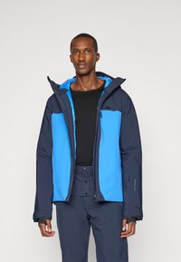 Kék és tengerészkék sídzseki, cipzáras zsebekkel, kapucnival és kontrasztos panelekből. Simább felület, részletes varrással és logóval a mellkason.