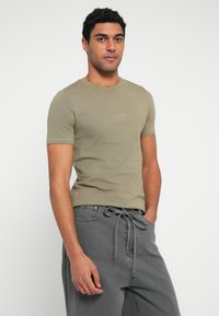 Λαδί κοντομάνικη μπλούζα με στρογγυλή λαιμόκοψη και διακριτικό λογότυπο. Συνδυασμένη με φαρδιά γκρι τζιν παντελόνια με δέσιμο στη μέση.