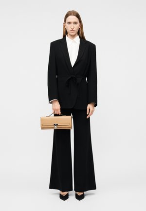 Frau trägt einen schwarzen maßgeschneiderten Anzug mit weit geschnittenen Hosen und einem weißen Hemd, hält eine beigefarbene rechteckige Handtasche mit oberem Griff.