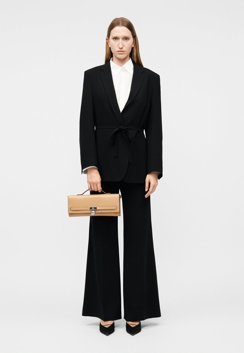 Γυναίκα που φοράει μαύρο κοστούμι ραμμένο στα μέτρα της με παντελόνι φαρδύ και λευκό πουκάμισο, κρατώντας μια μπεζ ορθογώνια τσάντα με πάνω χερούλι.