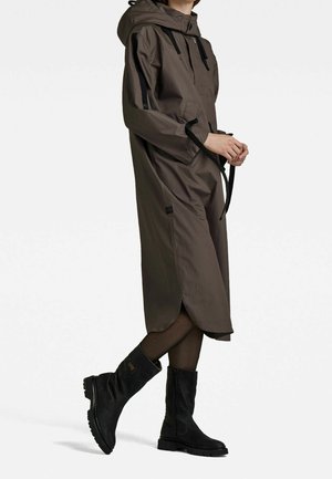 Gabardina impermeable marrón con capucha, detalles en negro, cremallera frontal y bolsillos laterales, combinada con botas negras hasta la rodilla.