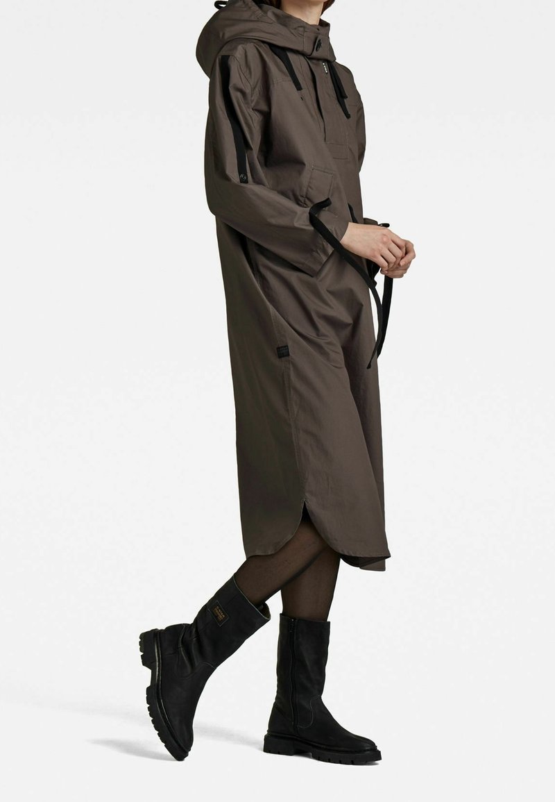 Brązowy wodoodporny płaszcz przeciwdeszczowy z kapturem, ozdobiony czarnymi akcentami, z przednim zamkiem błyskawicznym i bocznymi kieszeniami, w zestawieniu z czarnymi wysokimi kozakami do kolan.