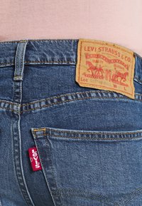Acercamiento al bolsillo trasero de unos vaqueros Levi's azules con la etiqueta roja de Levi's y un parche de cuero de Levi Strauss & Co. en la cintura, llevados sobre una camisa rosa.