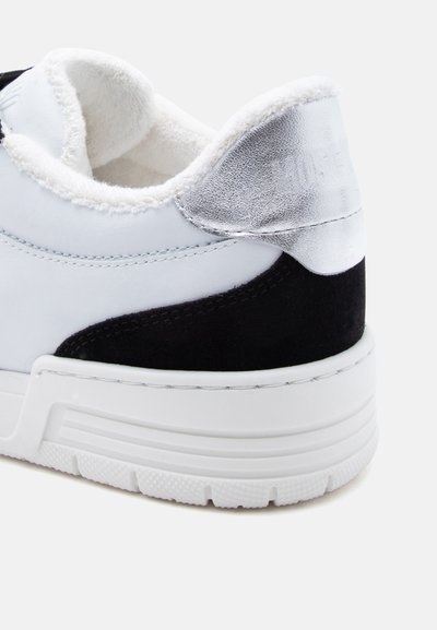 Λευκό τακούνι αθλητικού παπουτσιού με μαύρη λεπτομέρεια από σούεντ, γυαλιστερή ασημένια απόχρωση, παχύ λευκό πάτο και απαλή λευκή επένδυση ορατή στον γιακά.
