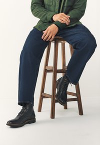 Schwarze Leder-Stiefeletten mit Brogue-Verzierung, getragen zu dunkelblauen Jeans und kombiniert mit einer grünen Jacke, sitzend auf einem hölzernen Hocker.