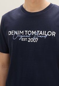 Camiseta de algodón azul marino con la inscripción "DENIM TOM TAILOR" en texto blanco y azul claro, cuello redondo y mangas cortas.