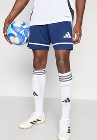 Blaue Sportshorts mit weißen Streifen, kombiniert mit weißen Schienbeinschonern mit schwarzem Muster. Weiße Fußballschuhe mit schwarzen Akzenten.