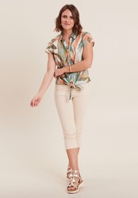 Korte mouwen blouse met een knoop aan de voorkant, voorzien van een meerkleurig bladerenpatroon in groen, oranje en perzik, gecombineerd met beige aansluitende broek en sandalen.