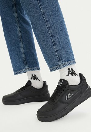Nogi w niebieskich dżinsach, białe skarpetki z czarnym logo oraz czarne sneakersy Kappa na jasnym tle.