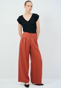 Zwarte V-hals top met opgerolde mouwen, gecombineerd met hoge taille, wijde oranje broek. Zwarte puntige hakken, gladde textuur, strakke lijnen.
