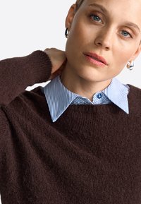Pull en maille marron avec une finition texturée, superposé sur une chemise à col rayée bleu et blanc. Présente des boucles d'oreilles en argent brillantes.