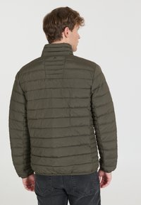 Veste matelassée vert olive avec des coutures horizontales, col haut et ourlet élastique. Matériau léger, adapté pour une utilisation en extérieur.