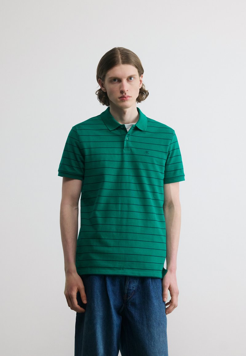 Jeune homme aux cheveux mi-longs portant un polo à rayures vertes et un jean bleu, debout devant un fond blanc uni.