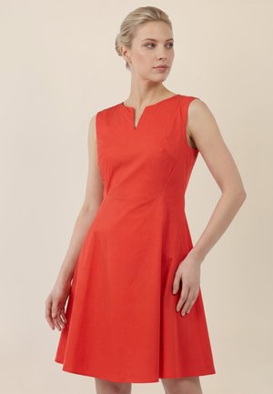 Frau mit blondem Haar, die ein ärmelloses, hellrotes Kleid mit kleinem V-Ausschnitt trägt, steht vor einem schlichten, hellen Hintergrund.