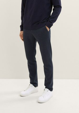Marineblaue Hose in schmaler Passform, aus einem strukturierten Stoff gefertigt. Kombiniert mit weißen Sneakern mit schwarzen Akzenten und flachen Schnürsenkeln.