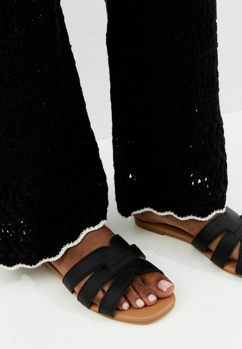 Pies con sandalias planas negras con tiras y suelas color crema, combinadas con pantalones negros texturizados con dobladillos blancos ondulados.