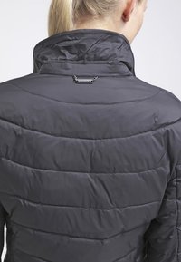 Blond kvinna i en svart vadderad jacka med hög krage sedd bakifrån mot en enfärgad bakgrund.
