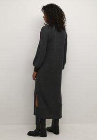 Robe en tricot gris foncé, longue et à la finition texturée, avec des fentes sur les côtés et de longues manches bouffantes, associée à des bottes noires montantes.