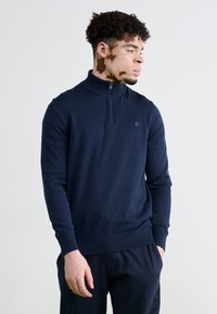 Tamno plavi pletena pulover s polu-zip ovratnikom, rebrastim manžetama i rubom; sadrži mali logo na prsima i glatku teksturu.