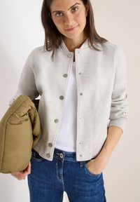 Hellgraue Bomberjacke aus glattem Stoff, mit Druckknöpfen, gerippten Bündchen und Kragen. Das Modell hält eine olivgrüne Daunenjacke.